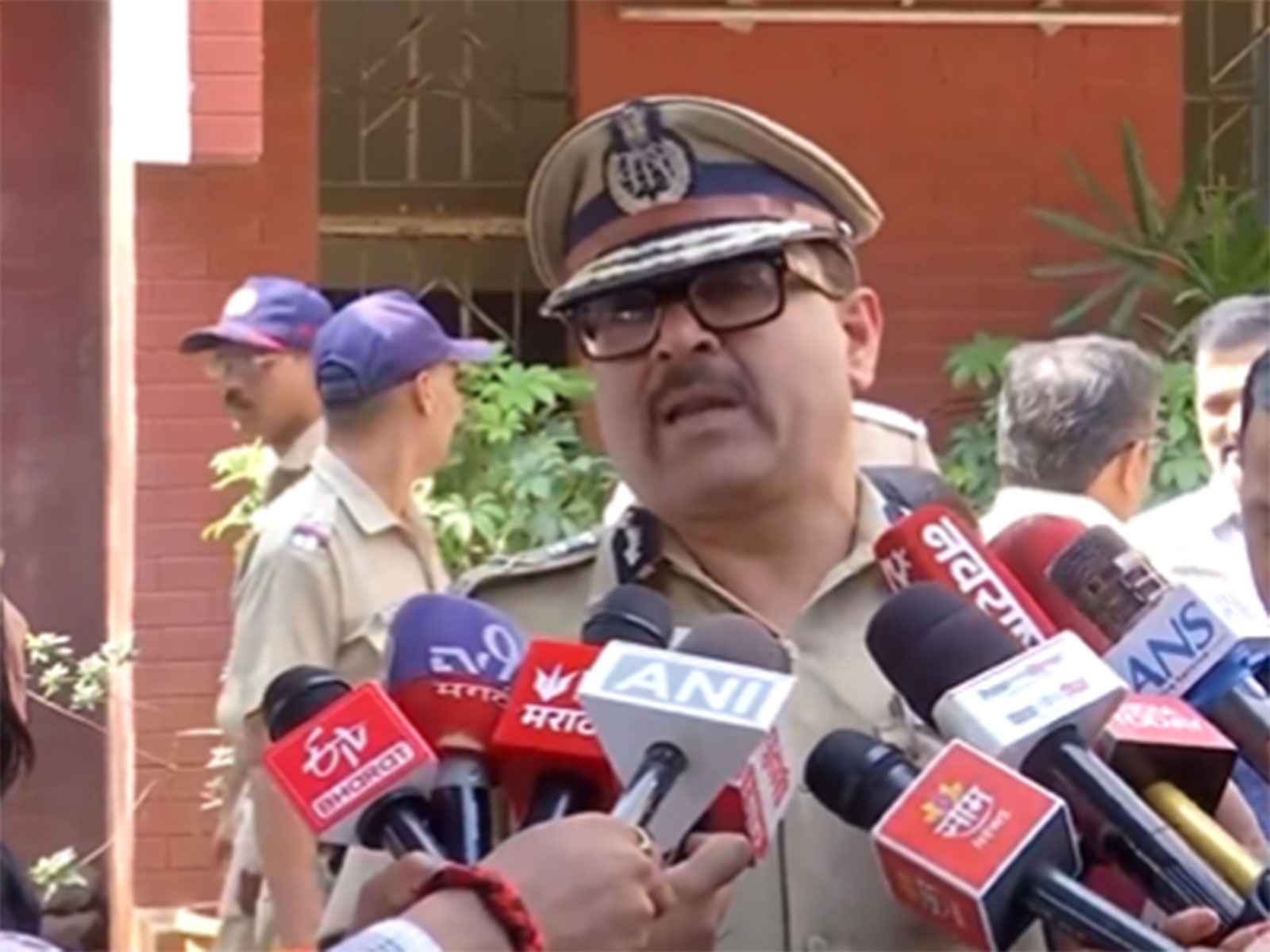 Amitesh Kumar briefing reporters in the field, surrounded by microphones, with police personnel behind him